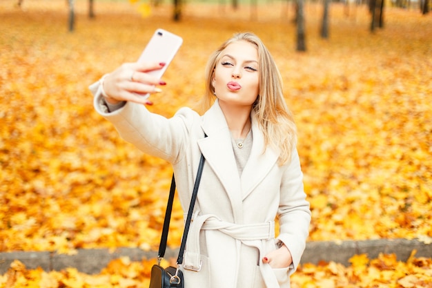 Stilvolles junges Mädchen, das selfie auf einem Smartphone am Herbsttag des gelben Laubes tut