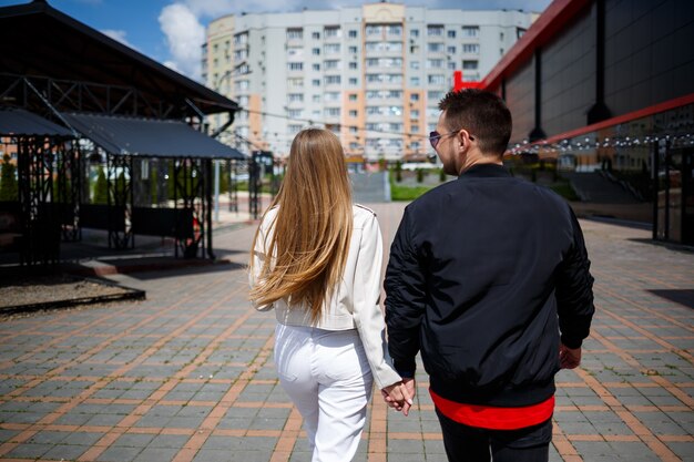 Stilvolles junges Mädchen mit blonden Haaren europäischen Aussehens und einem Mann in einer schwarzen Jacke. Urbaner Spaziergang eines verliebten Paares, moderne Mode im lässigen Stil. Glückliches Beziehungskonzept