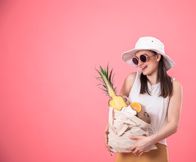 Stilvolles junges Mädchen mit weißem Hut und Sonnenbrille lächelt und hält eine Öko-Tasche mit exotischen Früchten Kopienraum.
