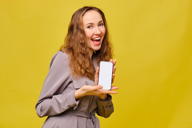 Stilvolles kaukasisches Mädchen in einem grauen Kleid auf gelbem Hintergrund. Er hält ein Smartphone in den Händen und lächelt.