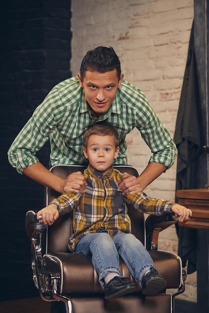 Stilvolles kleines Kind, das mit seinem jungen Vater im Hintergrund auf einem Stuhl im Friseursalon sitzt, herumalbern und Spaß haben