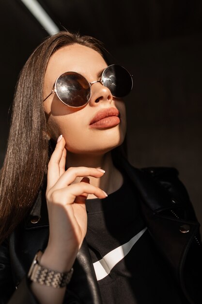 Stilvolles kreatives Porträt einer jungen Frau mit natürlichem Schönheitsgesicht und großen Lippen in schwarzer modischer Kleidung und runder Vintage-Sonnenbrille steht im Sonnenlicht und im Schatten auf dunklem Hintergrund