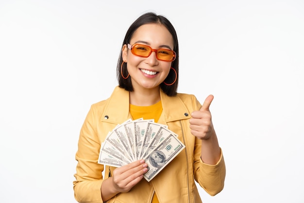 Stilvolles lächelndes asiatisches Mädchen, das Geld hält und Dollar zeigt und über weißem Hintergrund steht. Kopieren Sie Platz