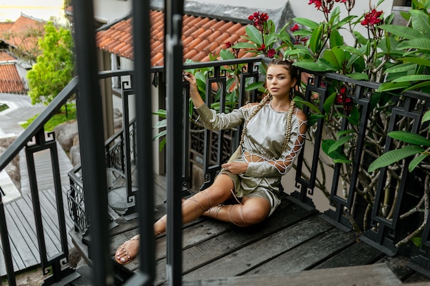 Stilvolles Mädchen im modernen Blick mit zwei langen Borten, die auf hölzerner Treppe nahe ihrem Haus sitzen