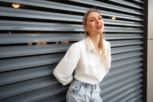 Stilvolles Mädchen in Jeans und weißer Baumwollbluse posiert in der Nähe der grauen Wand