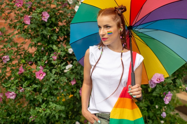 Stilvolles Mädchen mit lgbt Flagge auf ihrem Gesicht, das mit Regenbogenregenschirm aufwirft