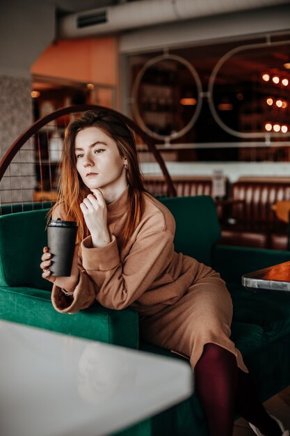 Stilvolles Mädchen sitzt in einem Café am Tisch und trinkt Kaffee. Kaffee zum Mitnehmen im Pappbecher. Frau mit roten Haaren in einem beigen warmen Anzug in gemütlicher Atmosphäre. Modernes Interieur. Ruhiger und angenehmer Zeitvertreib.