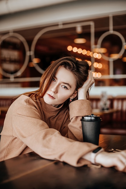 Stilvolles Mädchen sitzt in einem Café am Tisch und trinkt Kaffee. Kaffee zum Mitnehmen im Pappbecher. Frau mit roten Haaren in einem beigen warmen Anzug in gemütlicher Atmosphäre. Modernes Interieur. Ruhiger und angenehmer Zeitvertreib.