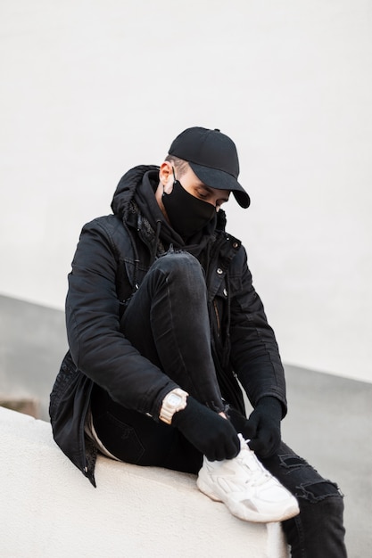 Stilvolles Mannmodell mit medizinischer Maske und Mütze in modischer urbaner Kleidung mit Jacke. Hoodie und Turnschuhe sitzt und bindet seine Schnürsenkel auf der Straße