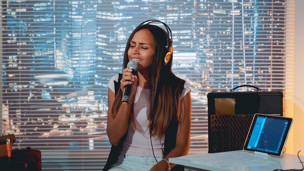 Stilvolles Mixed-Race-Mädchen, das bei der Probe im Heimstudio mit Wolkenkratzern im Hintergrund in das Mikrofon singt.