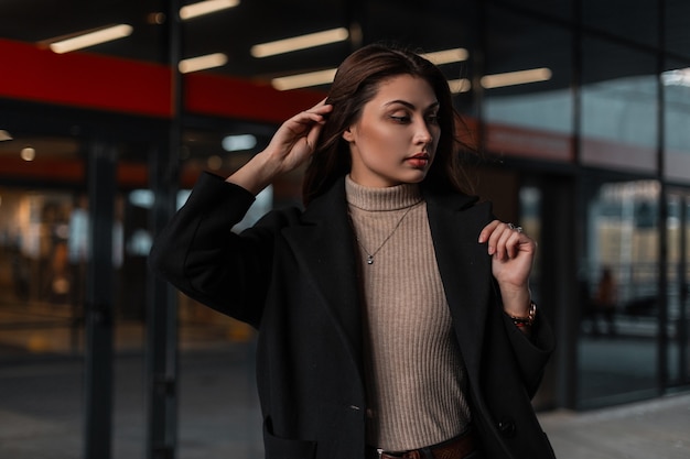 Stilvolles Modell hübsche junge Frau in einem modischen schwarzen Mantel in einem beigen Hemd posiert in der Nähe eines Vintage-Glasgebäudes in der Stadt. Nettes Modemädchen in der eleganten Kleidung draußen. Trendige Kleidung für Damen.