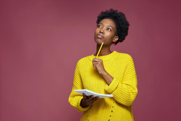Stilvolles nachdenkliches afroamerikanisches Mädchen, das über die Idee nachdenkt, ins Notizbuch zu schreiben und über das Geschäft nachzudenken