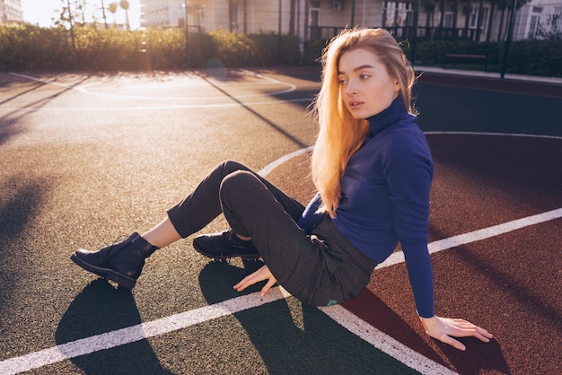 Stilvolles schönes blondes Modellmädchen im blauen Pullover sitzt in der Morgensonne auf dem Spielplatz