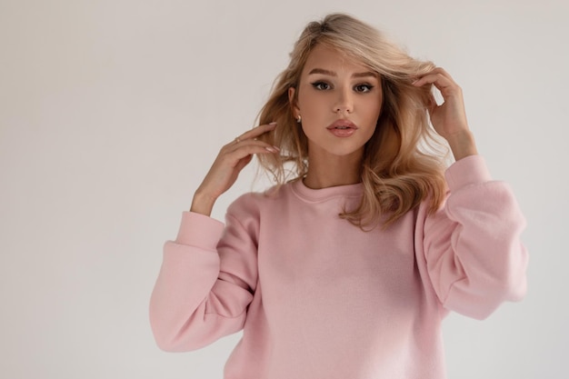 Stilvolles schönes blondes Modellmädchen in einem modischen rosa Pullover auf einem grauen Hintergrund im Studio betrachtet die Kamera