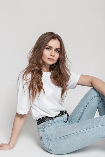 Stilvolles, schönes junges Mädchen mit Frisur in modischer Kleidung mit weißem T-Shirt und blauer Vintage-Jeans sitzt im Studio