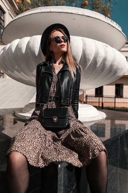 Stilvolles schönes junges Mädchenmodell in modischem Outfit mit Lederjacke und Vintage-Handtasche sitzt an einem sonnigen Tag auf der Straße. Weiblicher Sommerstil und Schönheit