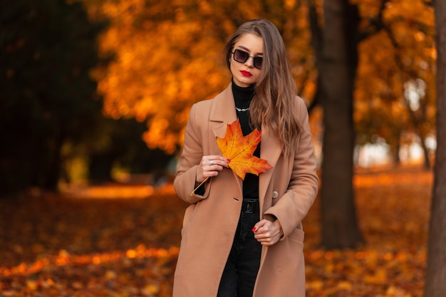 Stilvolles schönes Mädchen mit Sonnenbrille in modischer Kleidung und einem Mantel mit einem orange-roten Blatt geht in einem Herbstpark mit goldenem Laub spazieren