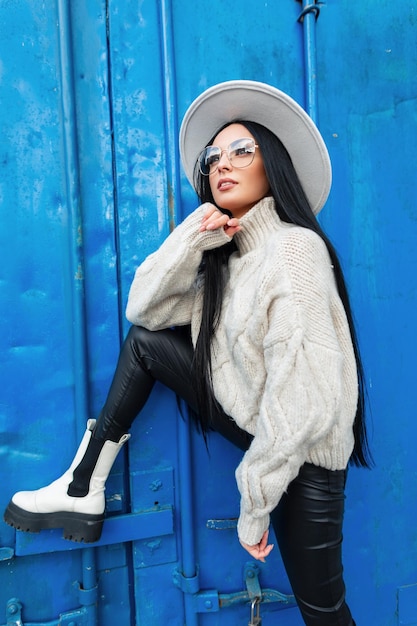 Stilvolles Schönheitsmode-Mädchenmodell mit trendiger Brille und Hut in weißen Strickpullovern, Lederhosen und Stiefeln posiert in der Nähe einer farbblauen Metallwand