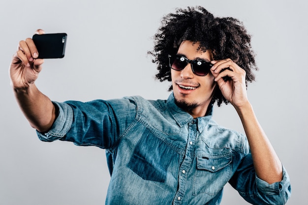 Stilvolles Selfie. Fröhlicher junger Afrikaner mit Sonnenbrille und Selfie im Stehen