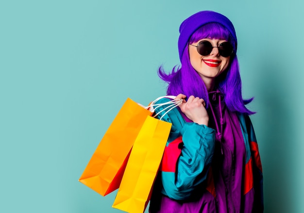 Stilvolles Shopaholic-Mädchen im Trainingsanzug und in der Sonnenbrille halten Einkaufstaschen an der blauen Wand