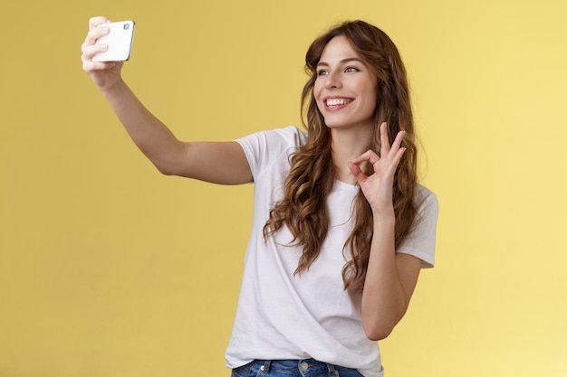 Stilvolles sorgloses städtisches selbstbewusstes lockiges Mädchen, das Video-Blog-Follower aufzeichnet, strecken den Arm aus und halten das Smartphone, das Selfie-Frontkamera zeigt, okay, ok Genehmigungsgeste gelber Hintergrund