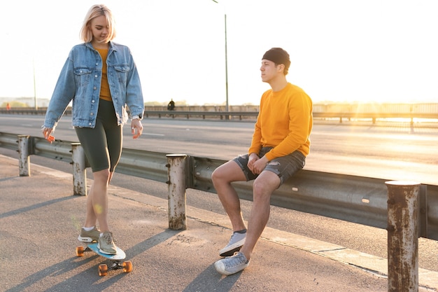 Stilvolles Teenager-Paar mit einem Longboard auf der Brücke während des Sonnenuntergangs