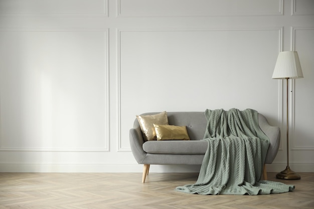Stilvolles Zimmerinterieur mit bequemer Sofa-Strickdecke und Lampe in der Nähe der weißen Wand Platz für Text