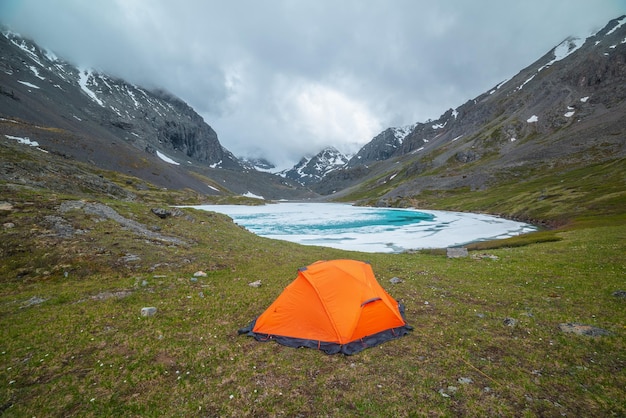 Stimmungsvolle Berglandschaft mit orangefarbenem Zelt in der Nähe eines zugefrorenen Alpensees und schneebedeckten Bergen. Fantastische Landschaft mit eisigem Bergsee vor dem Hintergrund schneebedeckter Berge in niedrigen Wolken. Zelt in der Nähe eines Eissees