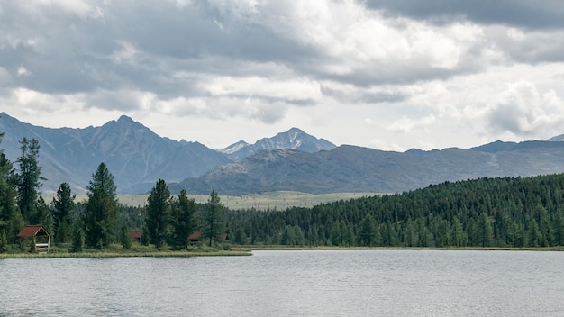 Stimmungsvoller Blick auf einen Bergsee mit Camping am Ufer in einem Nadelwald vor der Kulisse von Bergen und einem dramatisch bewölkten Himmel