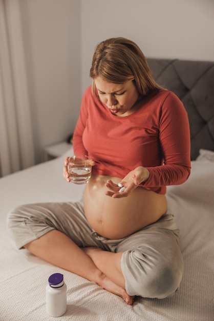 Stirnrunzelnde junge schwangere Frau, die sich unwohl fühlt, Aspirin oder Schmerzmittel auf einem Bett im Schlafzimmer bekommt.