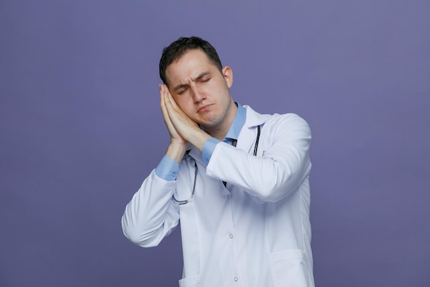 Stirnrunzelnder junger männlicher Arzt mit medizinischem Gewand und Stethoskop um den Hals, der eine Schlafgeste isoliert auf violettem Hintergrund macht
