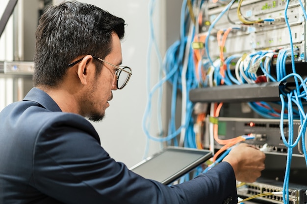 Stock Foto eines jungen Netzwerktechnikers, der Tablet hält, um Netzwerkkabel im Serverschrank im Netzwerkserverraum anzuschließen. IT-Ingenieur, der im Netzwerkserverraum arbeitet