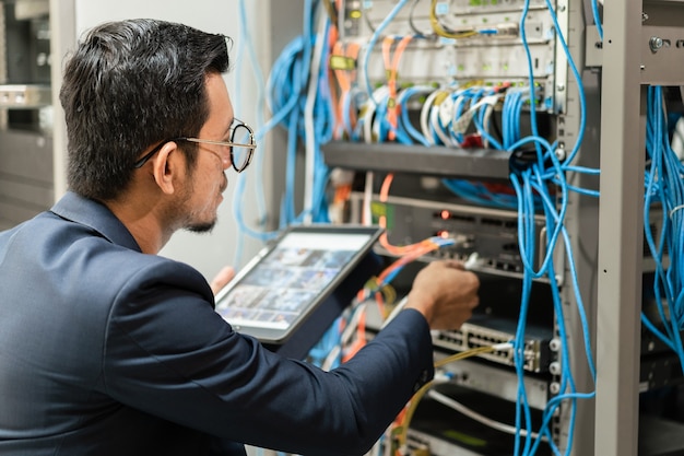 Stock Foto eines jungen Netzwerktechnikers, der Tablet hält, um Netzwerkkabel im Serverschrank im Netzwerkserverraum anzuschließen. IT-Ingenieur, der im Netzwerkserverraum arbeitet