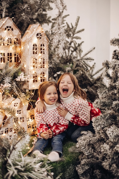 Stock Foto von fröhlichen Schwestern in roten und weißen Pullovern mit Weihnachtsschmuck lachend beim Kuscheln umgeben von Weihnachtsschmuck. Beleuchtete handgefertigte Häuser und Tannenzweige.