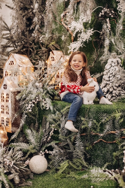 Stock Foto von schönen lächelnden brünetten Mädchen in rot-weißen Pullover und Jeans sitzen in Weihnachtsschmuck mit weißen Spielzeugkaninchen, Blick in die Kamera.