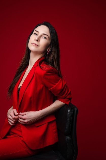 Stock photo retrato de una hermosa mujer de negocios segura de sí misma con el pelo oscuro en chaqueta roja sentado en una silla y mirando a la cámara. Aislar sobre fondo rojo brillante. Copie el espacio.