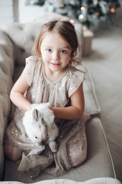 Stock photo retrato de niña linda en vestido festivo con conejito encantador en sus brazos sentado en el sofá. Árbol de Navidad decorado desenfocado en segundo plano.