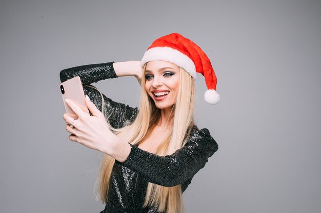 Stockfoto der attraktiven kaukasischen Frau in der roten Weihnachtsmütze und im grauen glänzenden Kleid, das Selfie über Smartphone macht