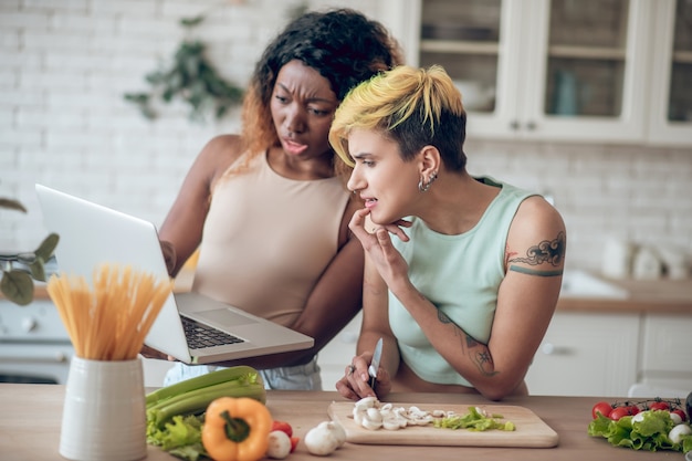 Störung. Emotionale junge Freundinnen mit fragender Grimasse, die am Laptop in der Küche am Morgen empört aussieht