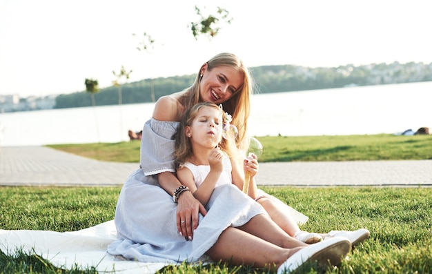 Stolze Mutter sieht Tochter an. Foto der jungen Mutter und ihrer Tochter, die gute Zeit auf dem grünen Gras mit See am Hintergrund haben.