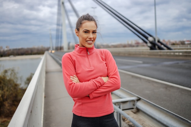 Stolze schöne lächelnde Sportlerin in Sportbekleidung, die auf der Brücke mit verschränkten Armen steht