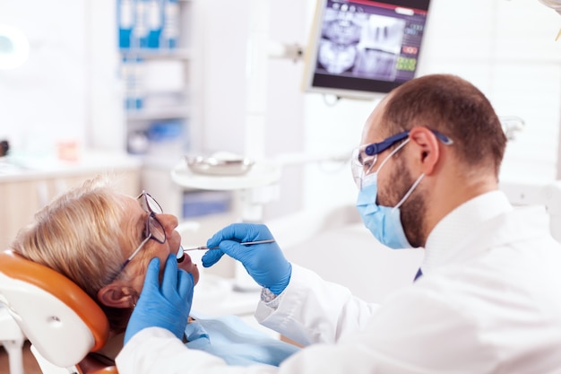 Stomatolog usando máscara facial durante a consulta de uma mulher sênior usando espelho em ângulo. Paciente idoso durante o exame médico com dentista no consultório odontológico com equipamento laranja.