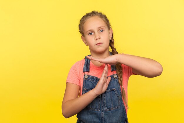 Stop-Limit-Geste Porträt eines müden, unglücklichen kleinen Mädchens mit Zopf in Denim-Overalls, das ein Timeout-Kind zeigt, das um Pause bittet, mit flehendem Ausdruck, Studioaufnahme isoliert auf gelbem Hintergrund