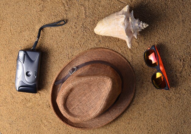 Strandzubehör liegt im Sand. Hut, Kamera, Sonnenbrille, Muschel. Das Konzept der Entspannung auf See. Die Sommerstrandsaison ist geöffnet! Draufsicht.