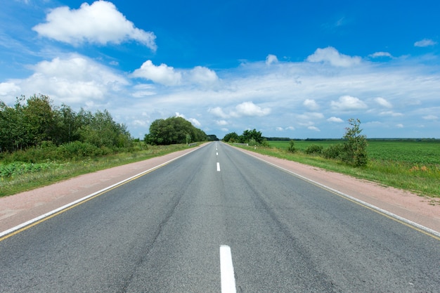 Straße durch das grüne Feld und die Wolken auf blauem Himmel am Sommertag