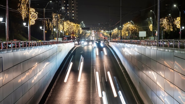 Straßenbild der Stadt bei Nacht, mehrere Autos, die sich auf der Straße innerhalb eines Tunnels bewegen, viel Weihnachtsbeleuchtung, Lichtspuren in Bukarest, Rumänien