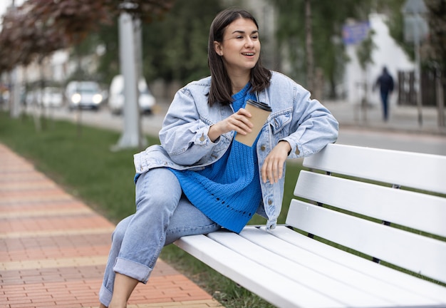 Straßenfoto einer attraktiven jungen Frau, die mit einem Kaffee in der Hand auf einer Bank sitzt.