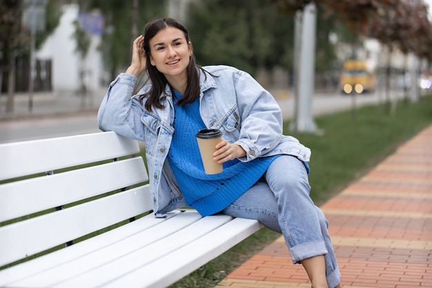 Straßenfoto einer attraktiven jungen Frau, die mit einem Kaffee in der Hand auf einer Bank sitzt