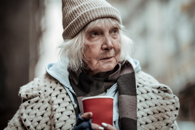 Straßenleben. Porträt einer alten traurigen armen Frau, die eine Tasse Tee hält