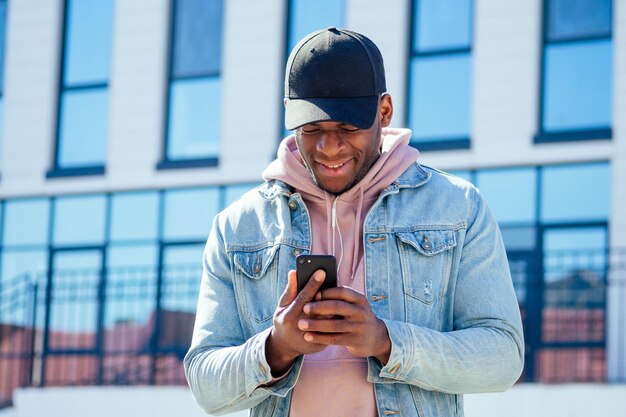 Straßenmodekonzept stilvoller gutaussehender afroamerikanischer mann in einer rosa hemdkapuze und einer jeansjacke und einer schwarzen kappe gegen die fenster des geschäftszentrums